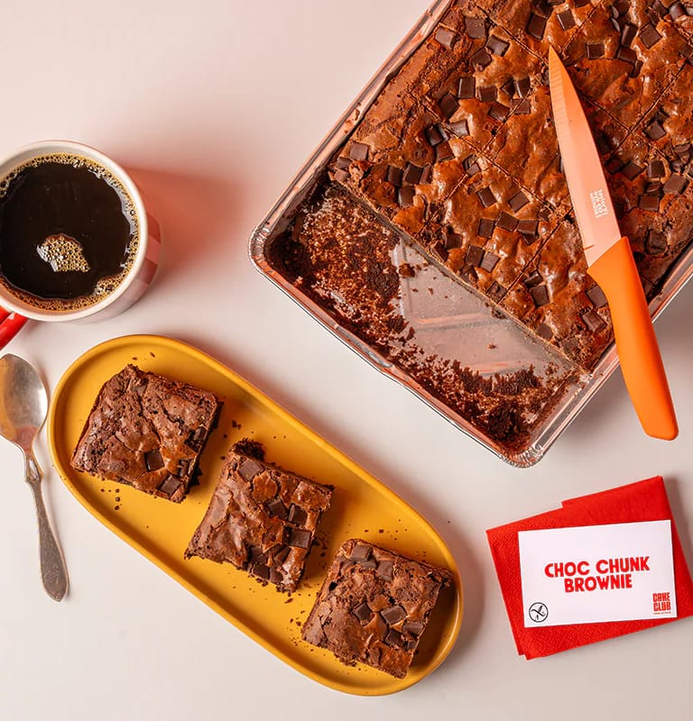 Three Choc Chunk Brownie slices cut and served on a yellow plate beside the remaining brownie tray and a coffee.